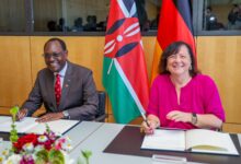 Kenyan and German officials sign a bilateral development cooperation agreement in Berlin with the Kenyan and German flags displayed behind them.