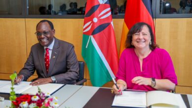Kenyan and German officials sign a bilateral development cooperation agreement in Berlin with the Kenyan and German flags displayed behind them.