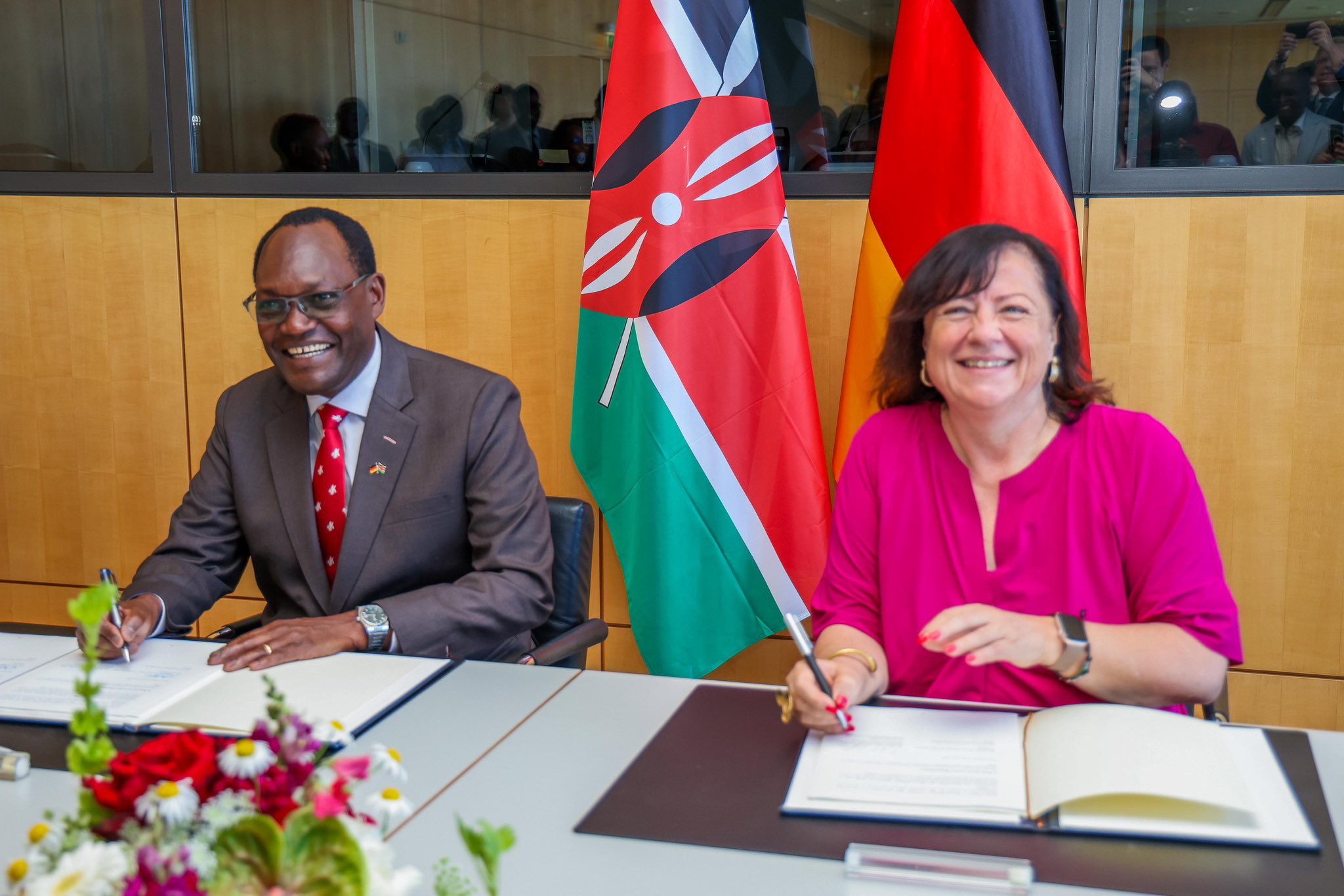 Kenyan and German officials sign a bilateral development cooperation agreement in Berlin with the Kenyan and German flags displayed behind them.