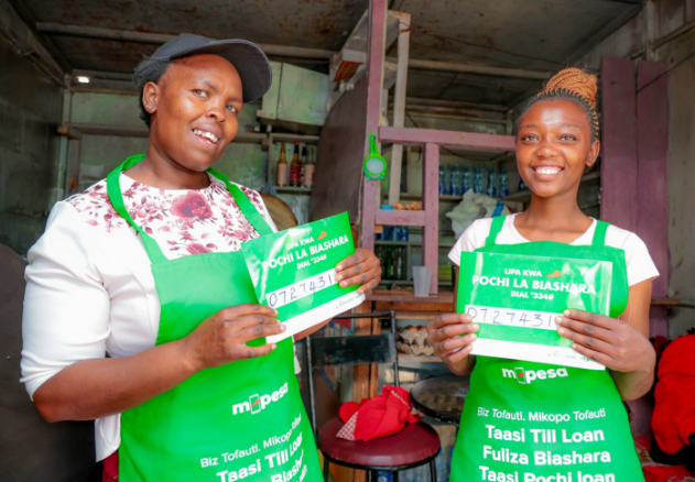 Two Kenyan women entrepreneurs holding Pochi la Biashara merchant signs inside a small retail shop.
