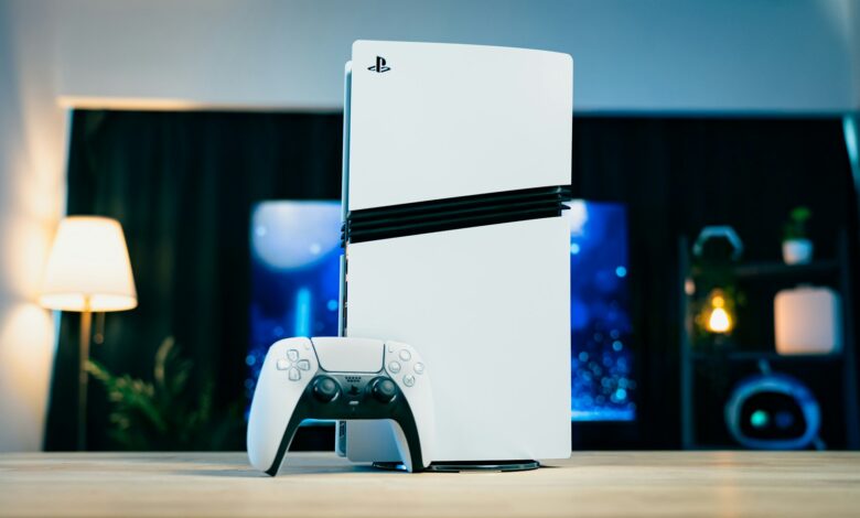 A white PlayStation 5 console standing beside a DualSense controller on a table in a modern gaming setup.
