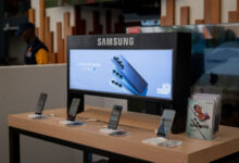 Samsung Galaxy S26 smartphones on a wooden retail display stand beneath a backlit Samsung Galaxy S26 Series banner featuring Galaxy AI.
