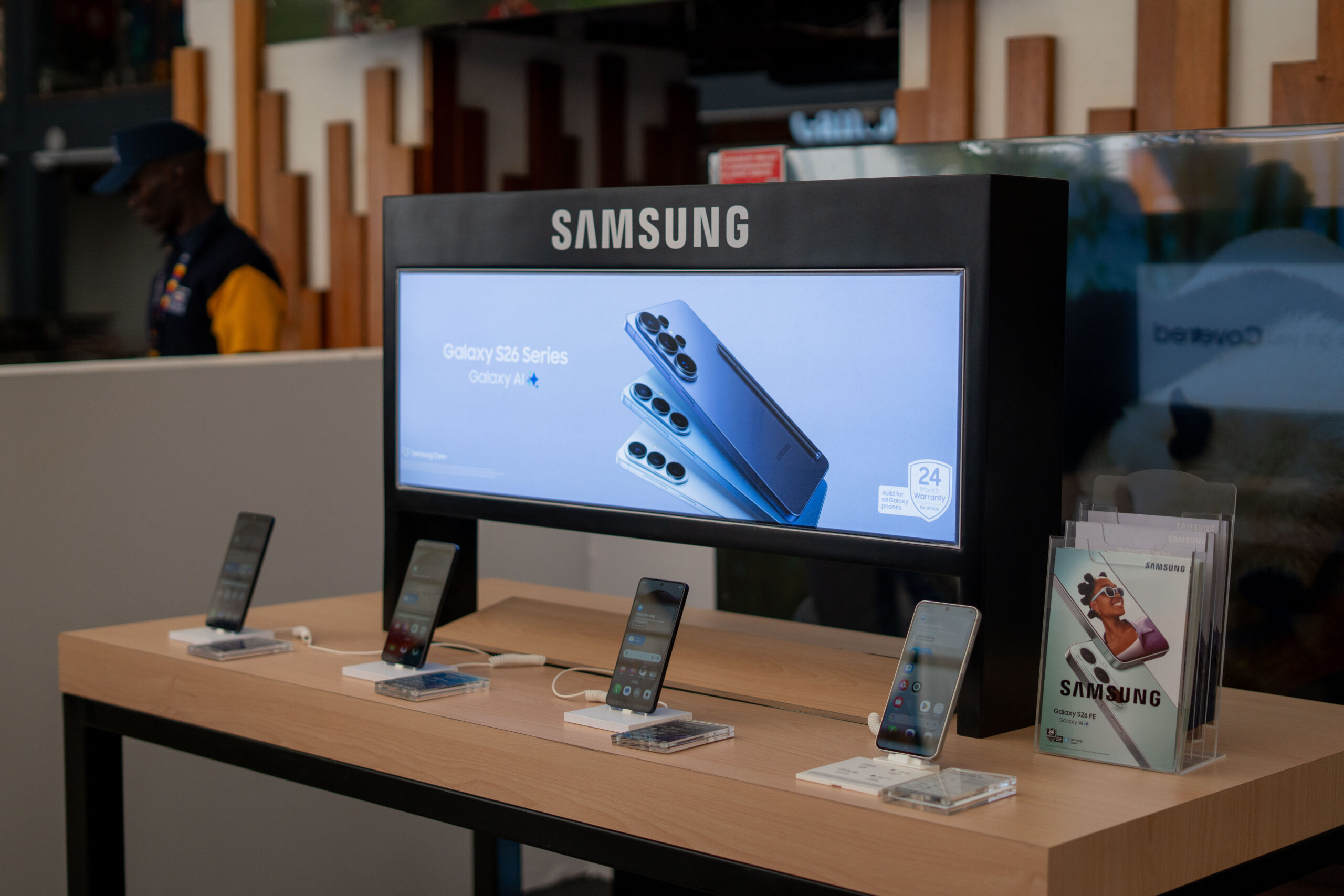 Samsung Galaxy S26 smartphones on a wooden retail display stand beneath a backlit Samsung Galaxy S26 Series banner featuring Galaxy AI.